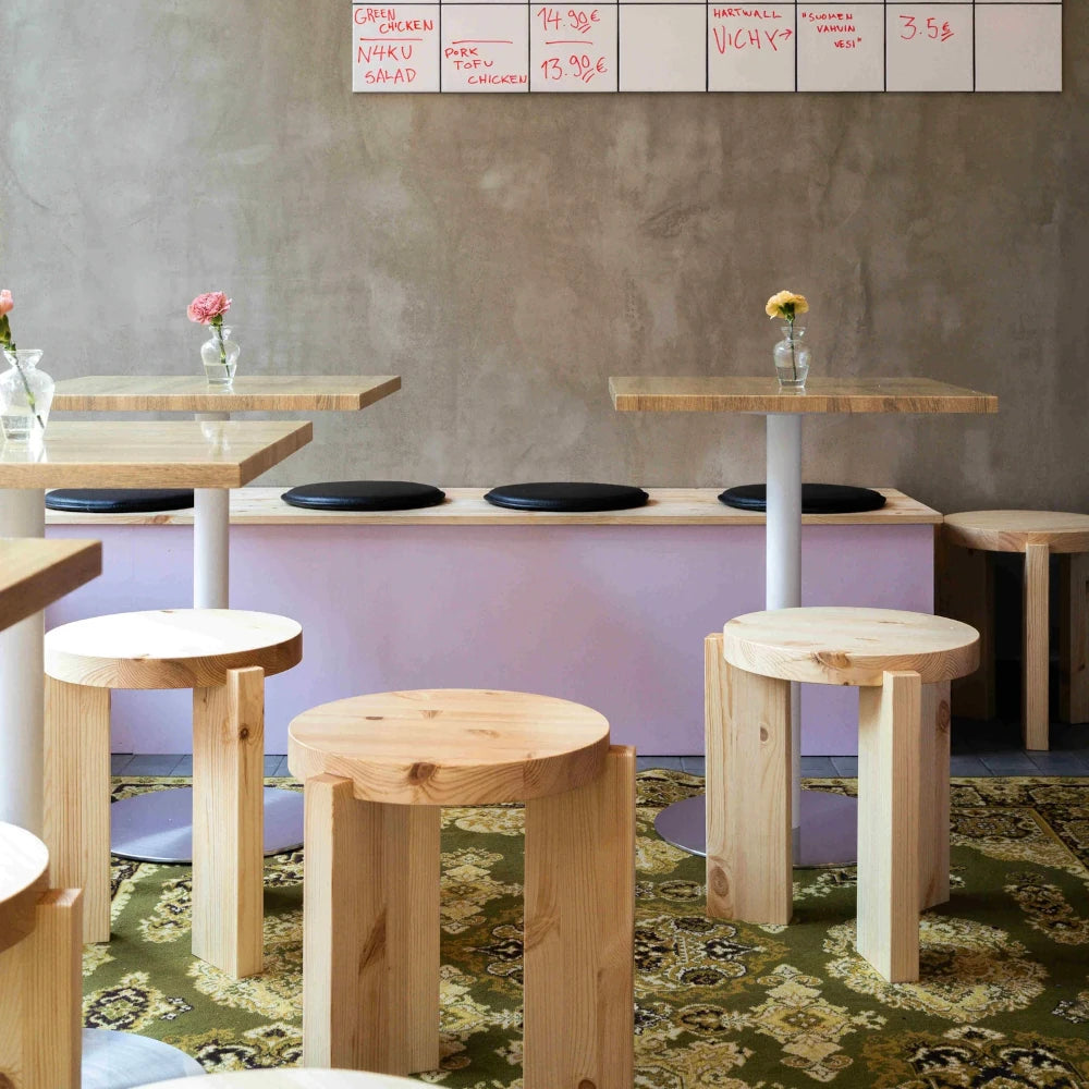 aburete bajo de madera natural con asiento redondo y tres patas de diseño geométrico, estilo nórdico y minimalista, en un espacio comercial contemporáneo.